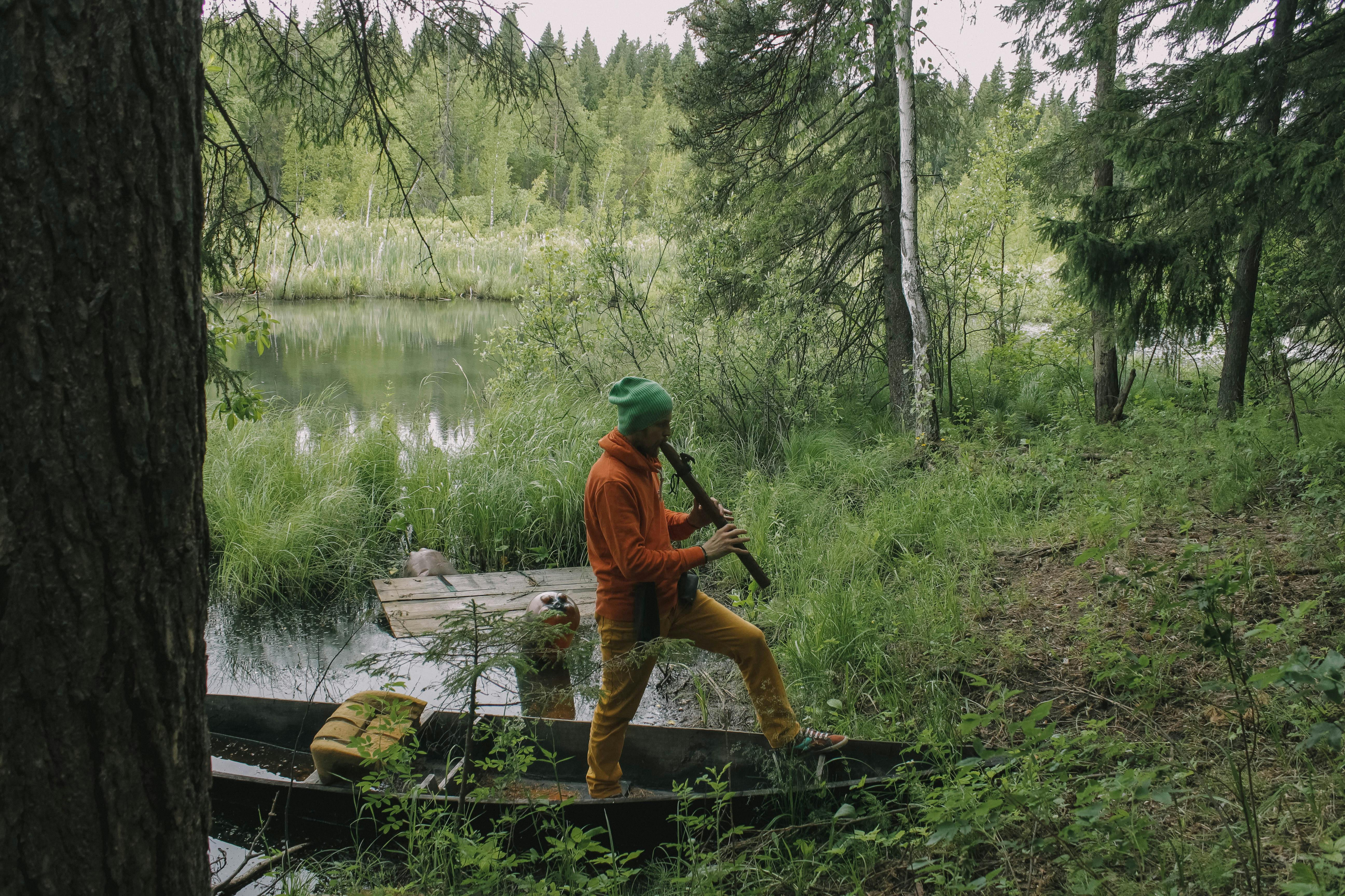 A musician plays a flute in a forest beside a river