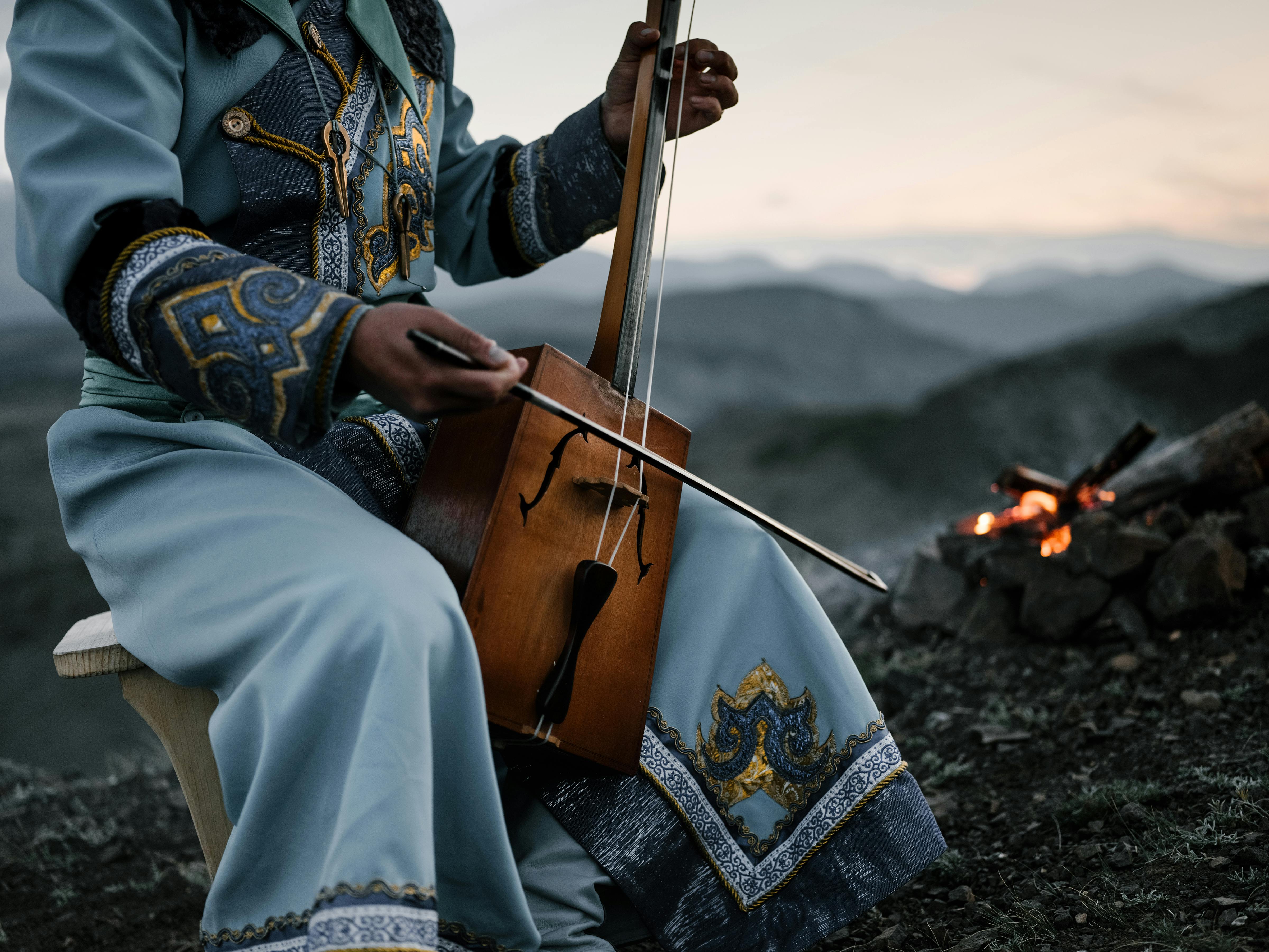 A musician in traditional dress plays a string instrument beside a campfire in the mountains at dusk