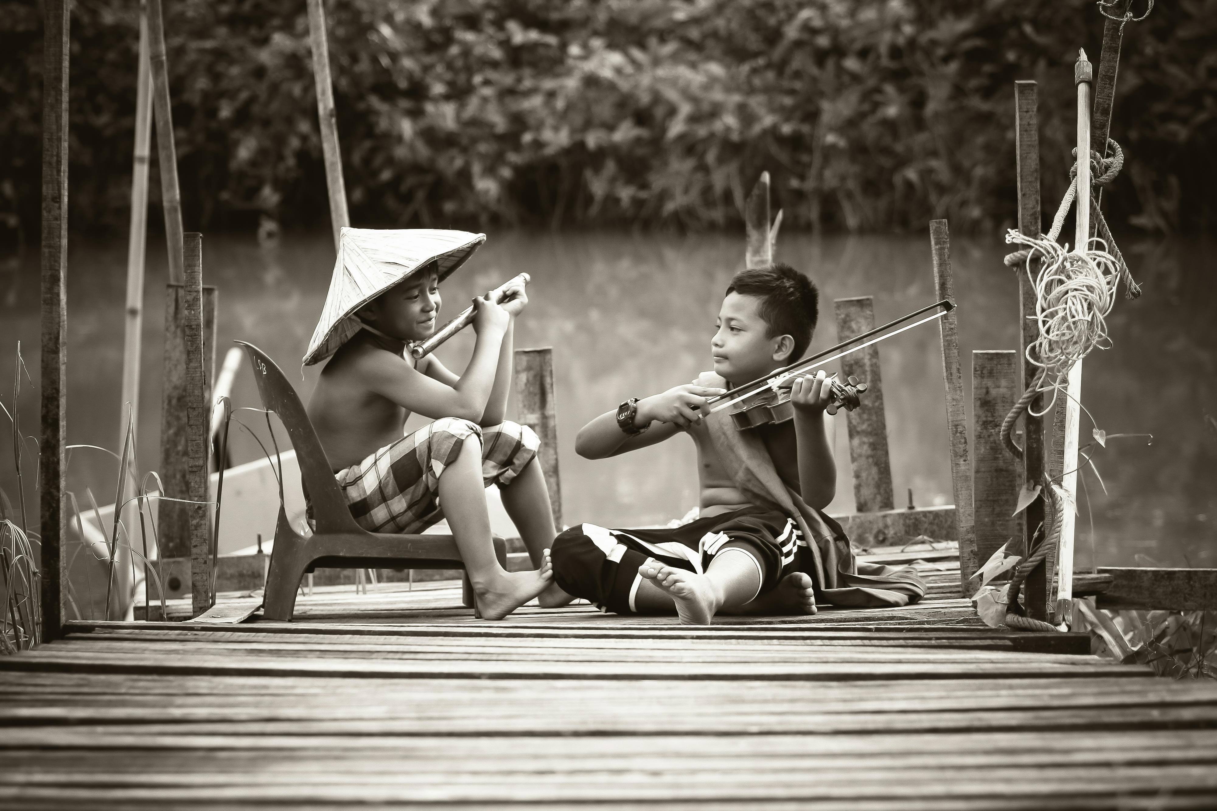 Two boys playing musical instruments together on a wooden dock by the water