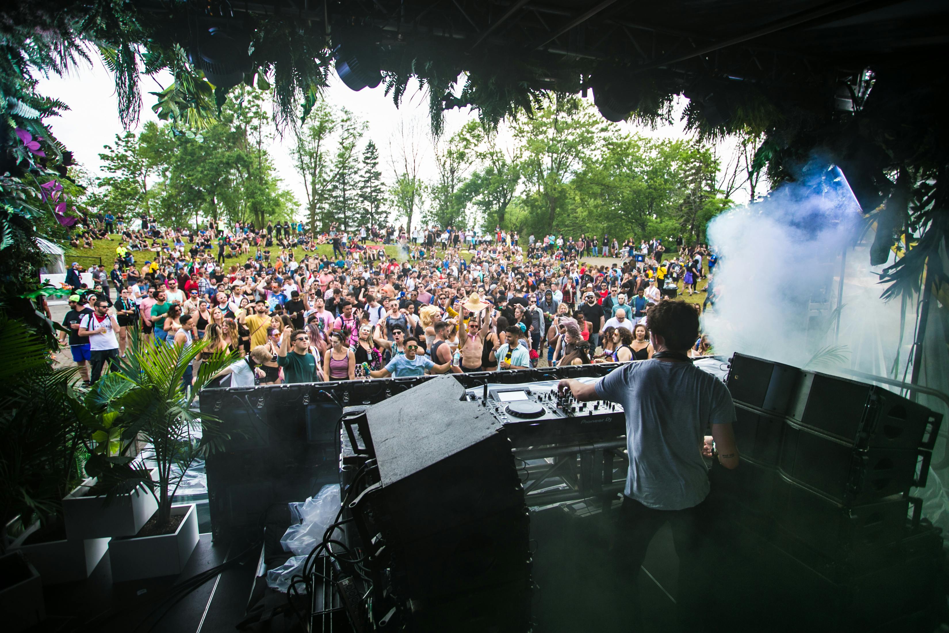 An artist on stage faces a crowd of thousands gathered under open sky surrounded by trees